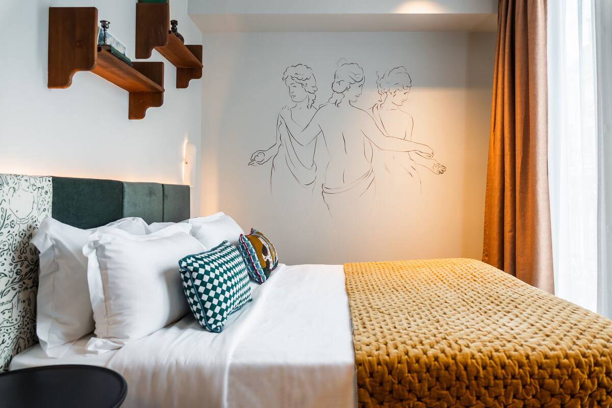 A neatly made bed with white and patterned pillows, a yellow textured blanket, and a wall with a line drawing of two figures.