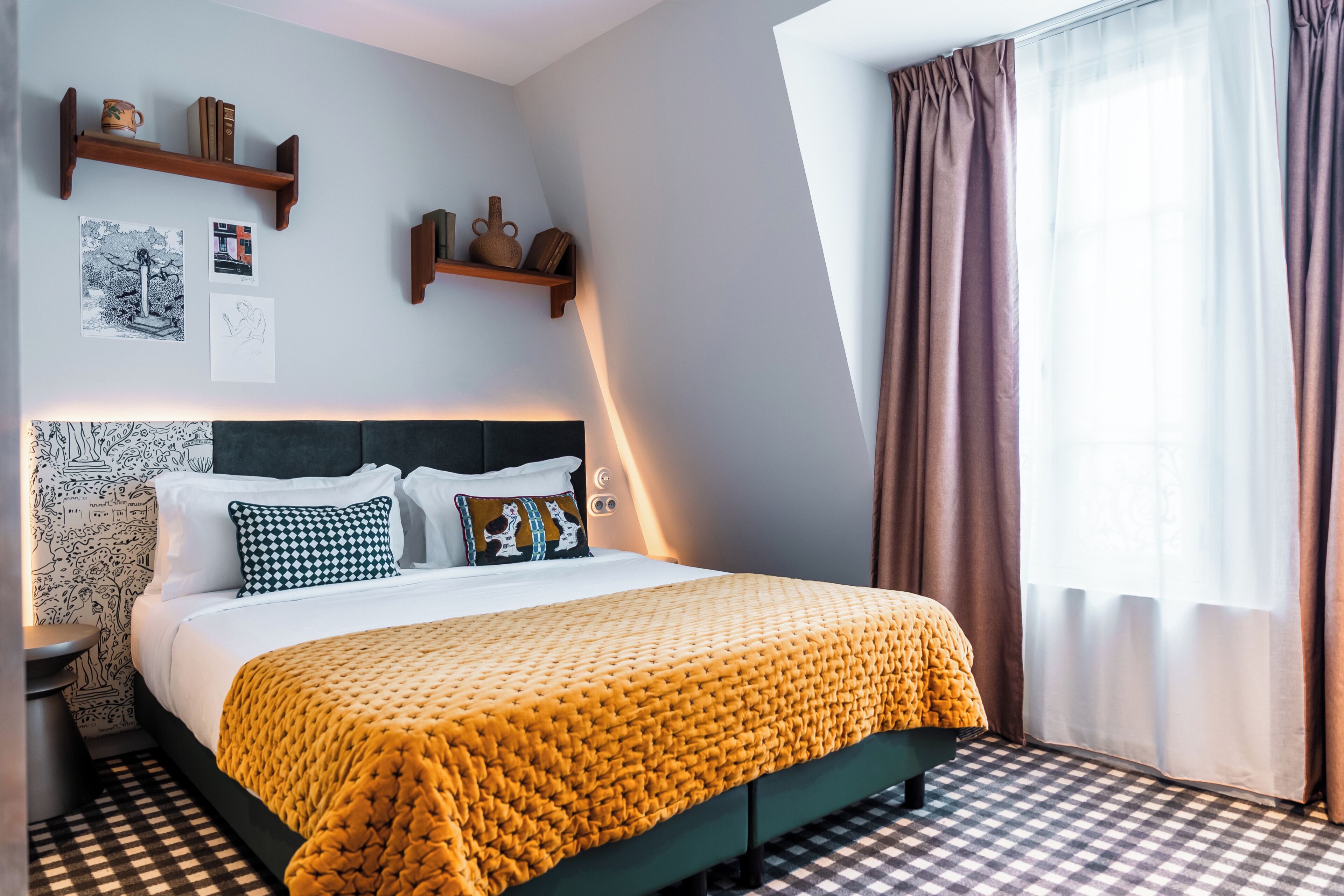 A neatly arranged hotel room with a checkered floor, a bed with a yellow blanket, a headboard with a patterned design, and a window with curtains.