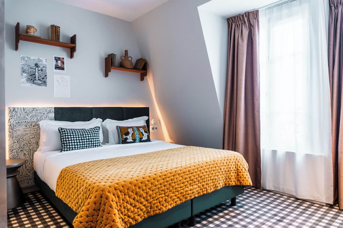 Ein ordentlich eingerichtetes Hotelzimmer mit kariertem Boden, einem Bett mit gelber Decke und einem ansprechend designten Kopfteil sowie einem Fenster mit Vorhängen.