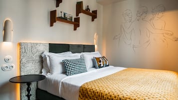 Ein ordentlich bezogenes Bett mit weißen Leinentüchern und einer gelben, strukturierten Decke, ein kleiner runder Tisch mit schwarzer Oberfläche und Holzregale mit Dekorationen.