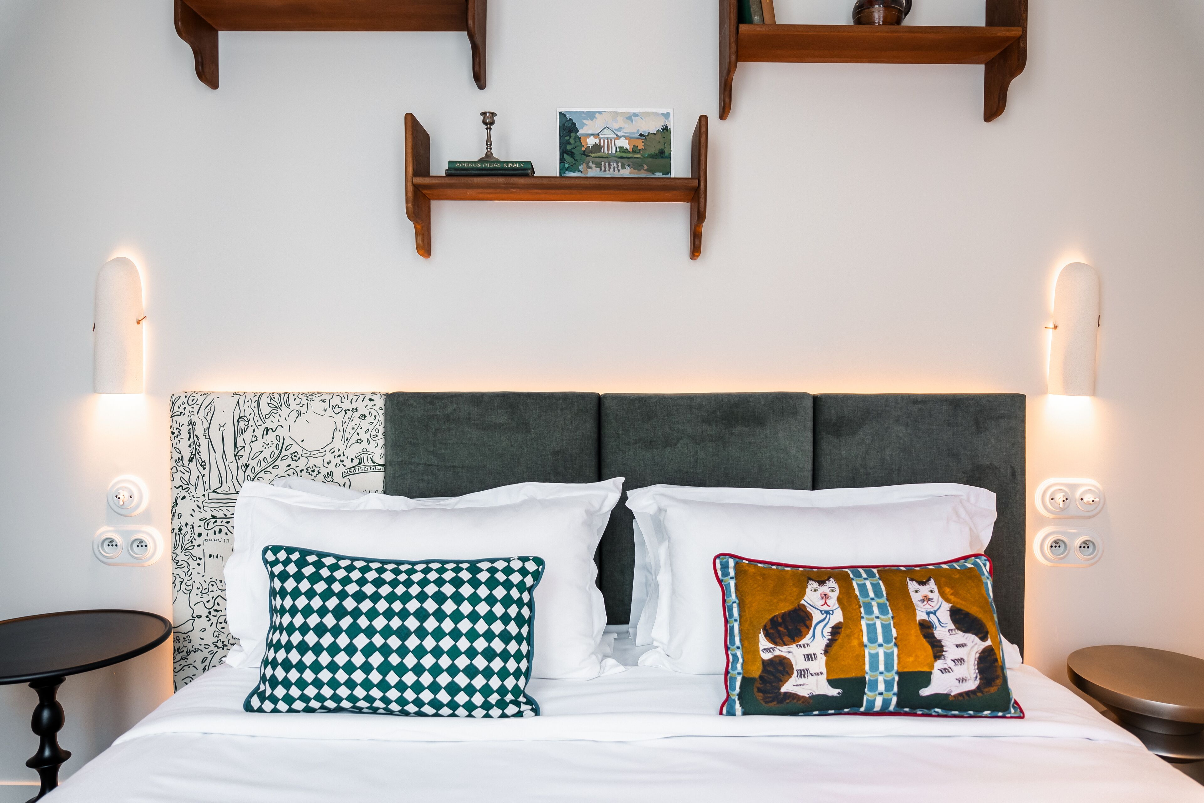 Una cama bien hecha con sábanas blancas, dos almohadas estampadas y un cabecero gris. Hay dos mesitas de noche, una a cada lado de la cama.