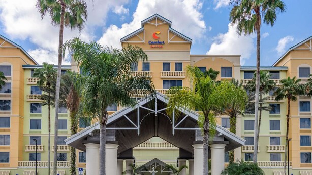 A hotel with a central entrance, palm trees, and a clear sky. - Comfort Suites Maingate East (Kissimmee)