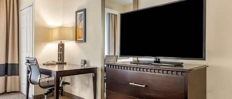 A hotel room with a desk, chair, TV, dresser, lamp, and a door.