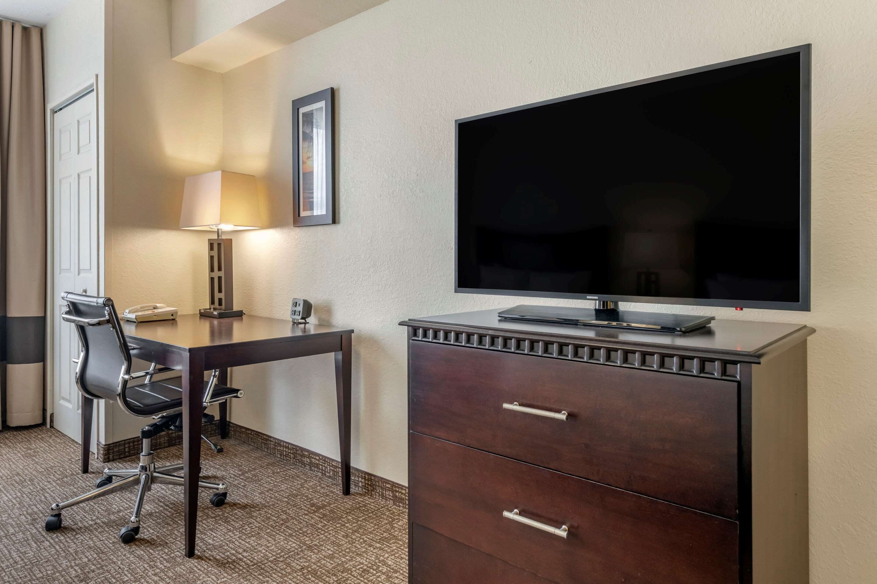 A hotel room with a desk, chair, TV, and a dresser.