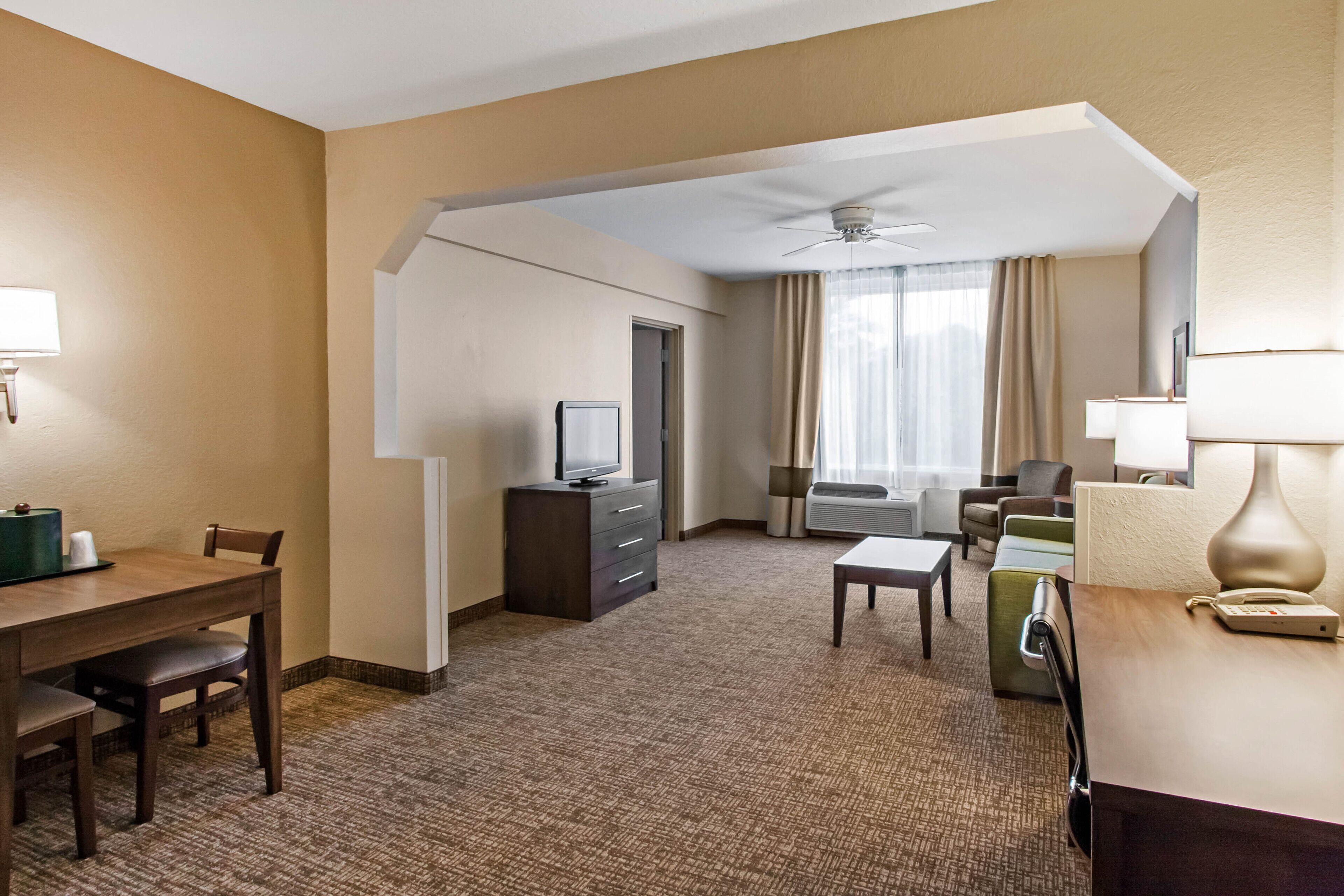 A hotel room with a dining area, a TV, a desk, a chair, a sofa, and a window with curtains.