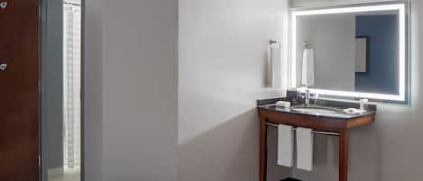 Une chambre d’hôtel avec un plancher en bois, un lit, une salle de bain avec un lavabo et un miroir, et une porte donnant sur une autre pièce.