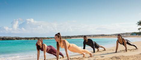 Four people doing push-ups on a sandy beach with a clear sky and calm sea in the background.