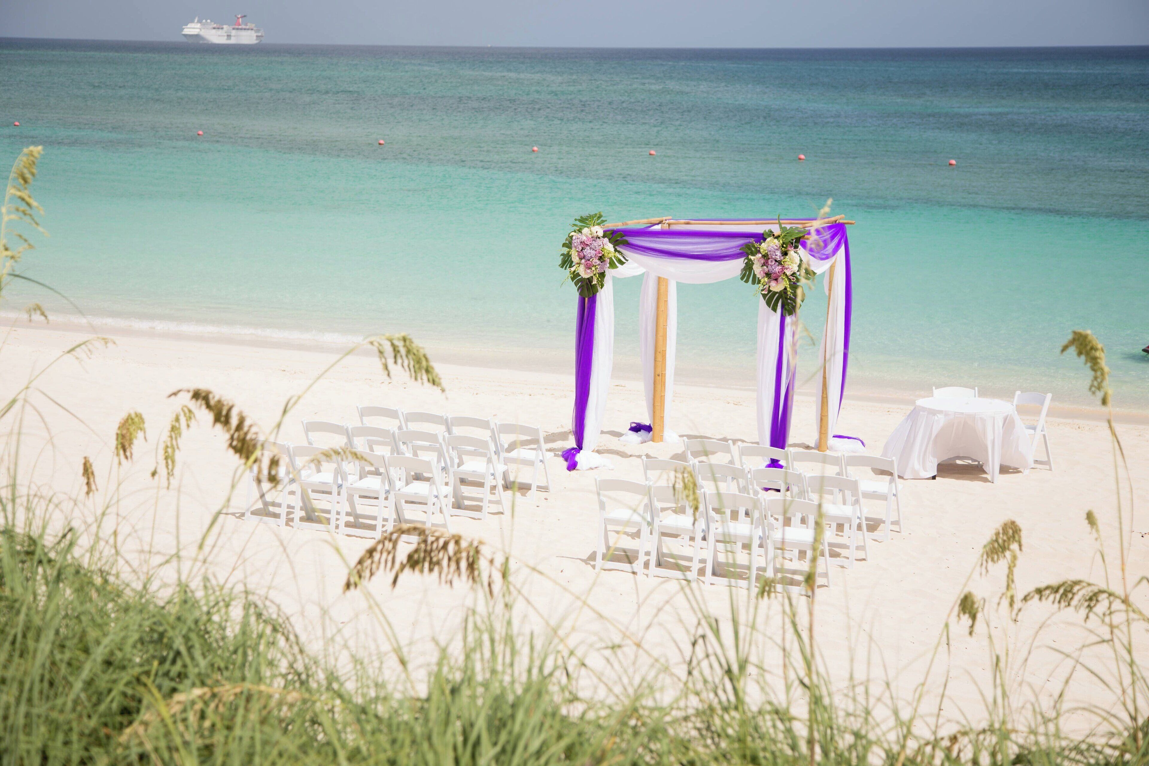 A beach wedding setup with white chairs and a table, a purple and white canopy, and a view of the ocean.