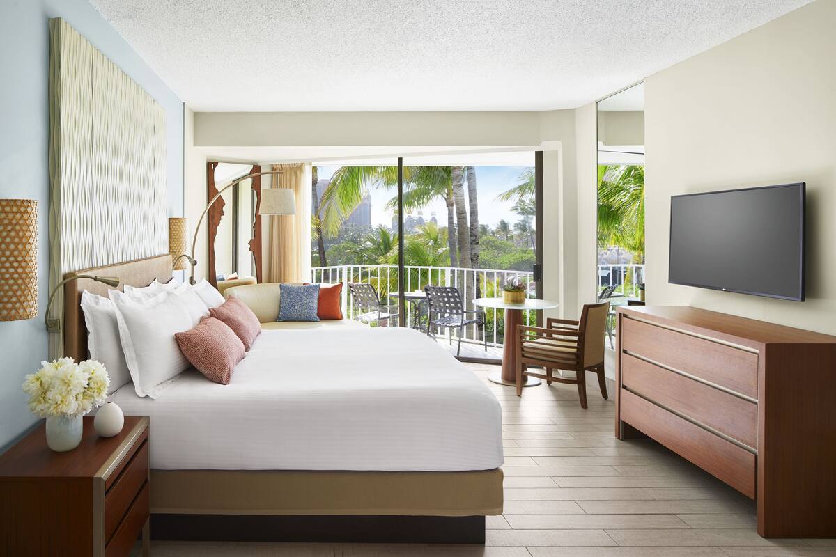 Ein Hotelzimmer mit einem großen Bett, einem Fernseher, einem Balkon mit Blick auf Palmen und einem Essbereich.