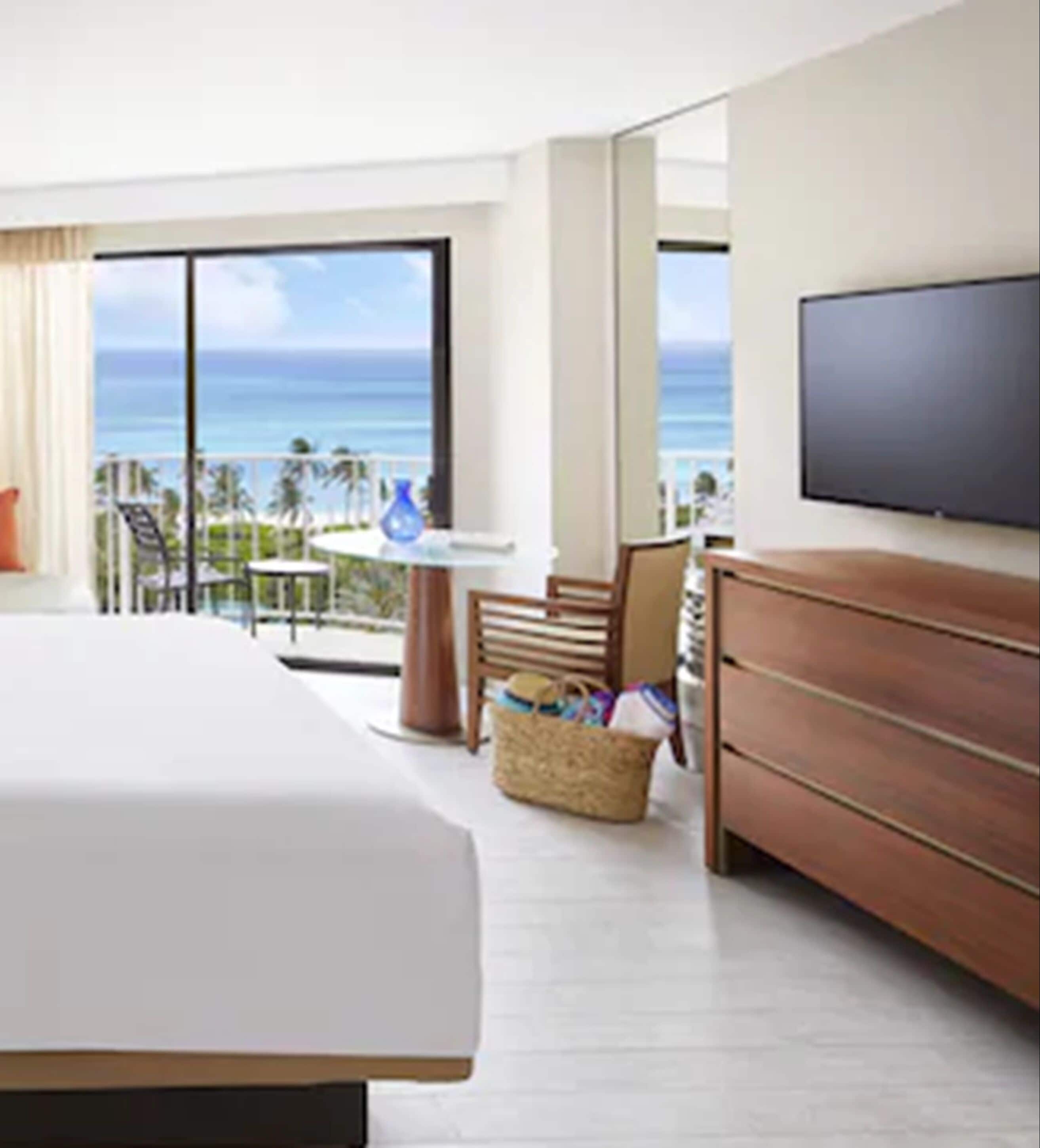 A hotel room with a large bed, a TV, a wooden dresser, a wicker basket, and a balcony with a view of the ocean.