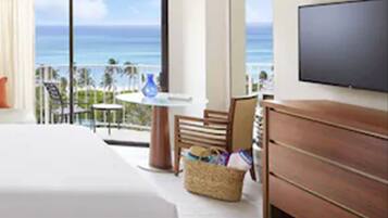 A hotel room with a large bed, a TV, a wooden dresser, a wicker basket, and a balcony with a view of the ocean.