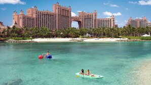 A resort with a large building featuring a distinctive archway, a beach, and clear turquoise waters. - The Coral at Atlantis (Paradise Island)