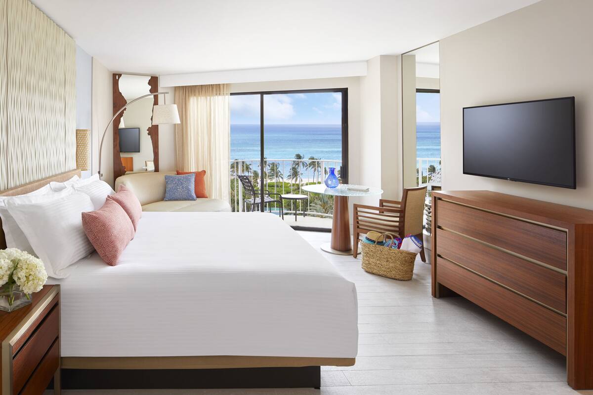 Ein Hotelzimmer mit einem großen Bett, einem Fernseher, einem Balkon mit Meerblick und einer Holzkommode.