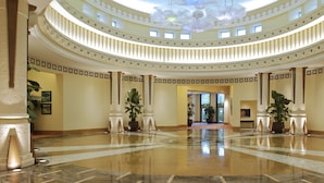 A grand, circular lobby with a high ceiling and a central chandelier. The lobby features a polished marble floor and is adorned with large potted plants.