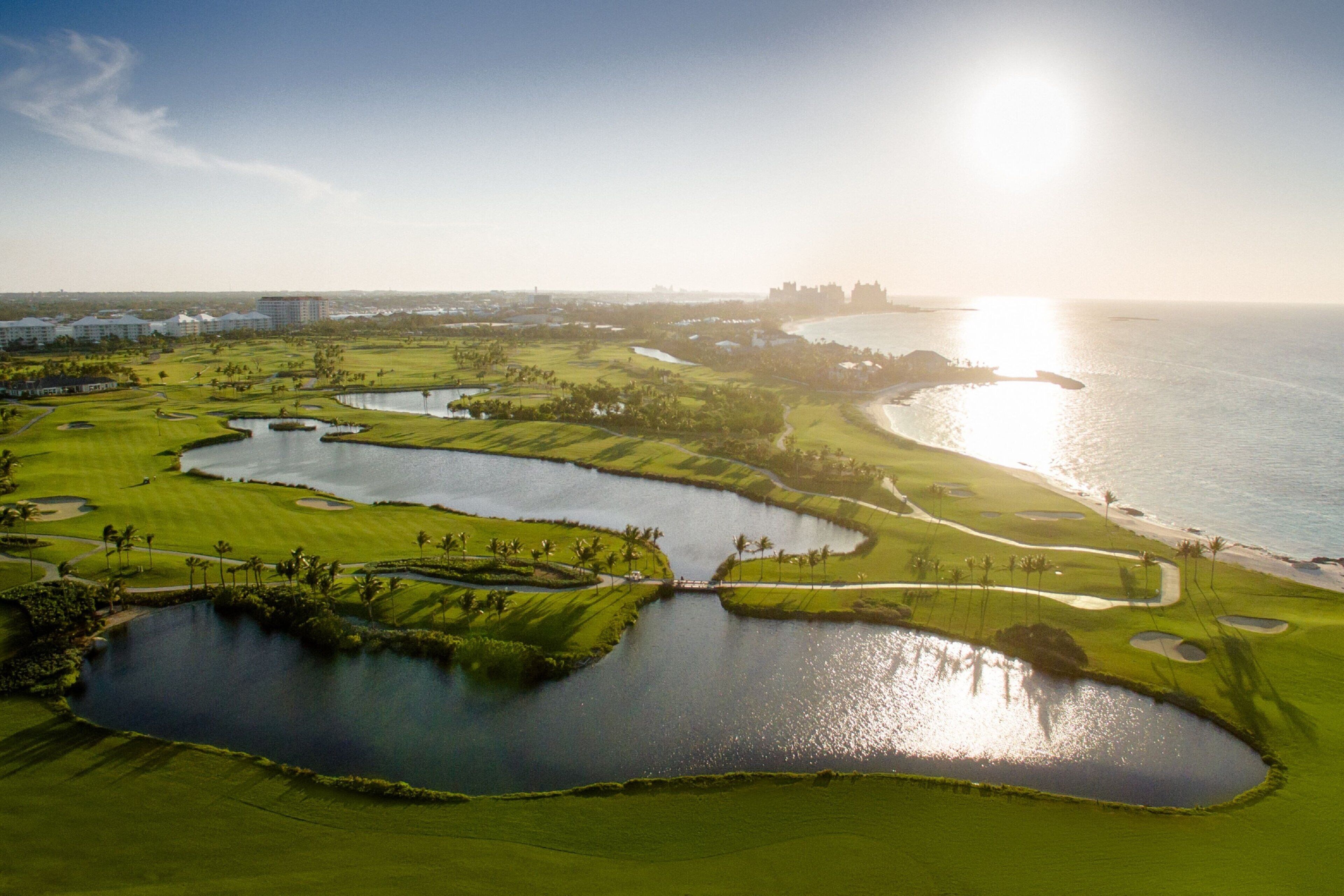 A golf course with multiple ponds and a beach area, surrounded by buildings and a coastline.