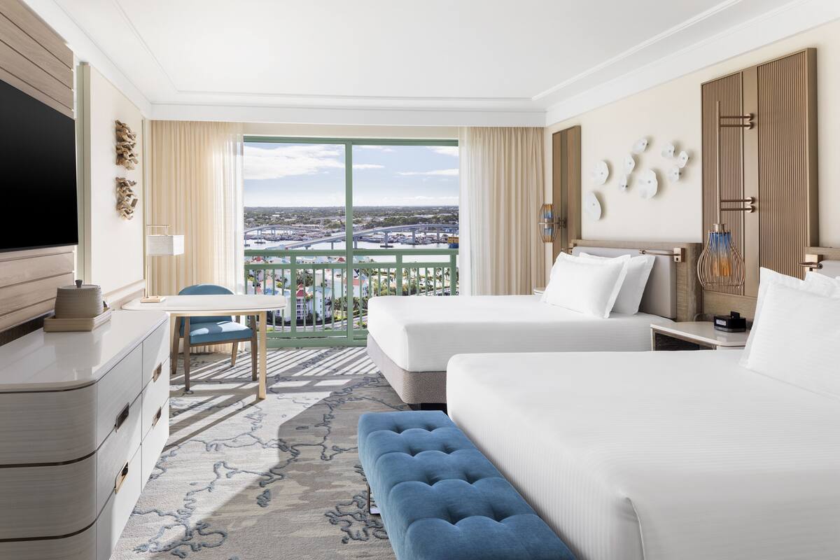 A hotel room with two beds, a TV, a desk, and a balcony with a city view.