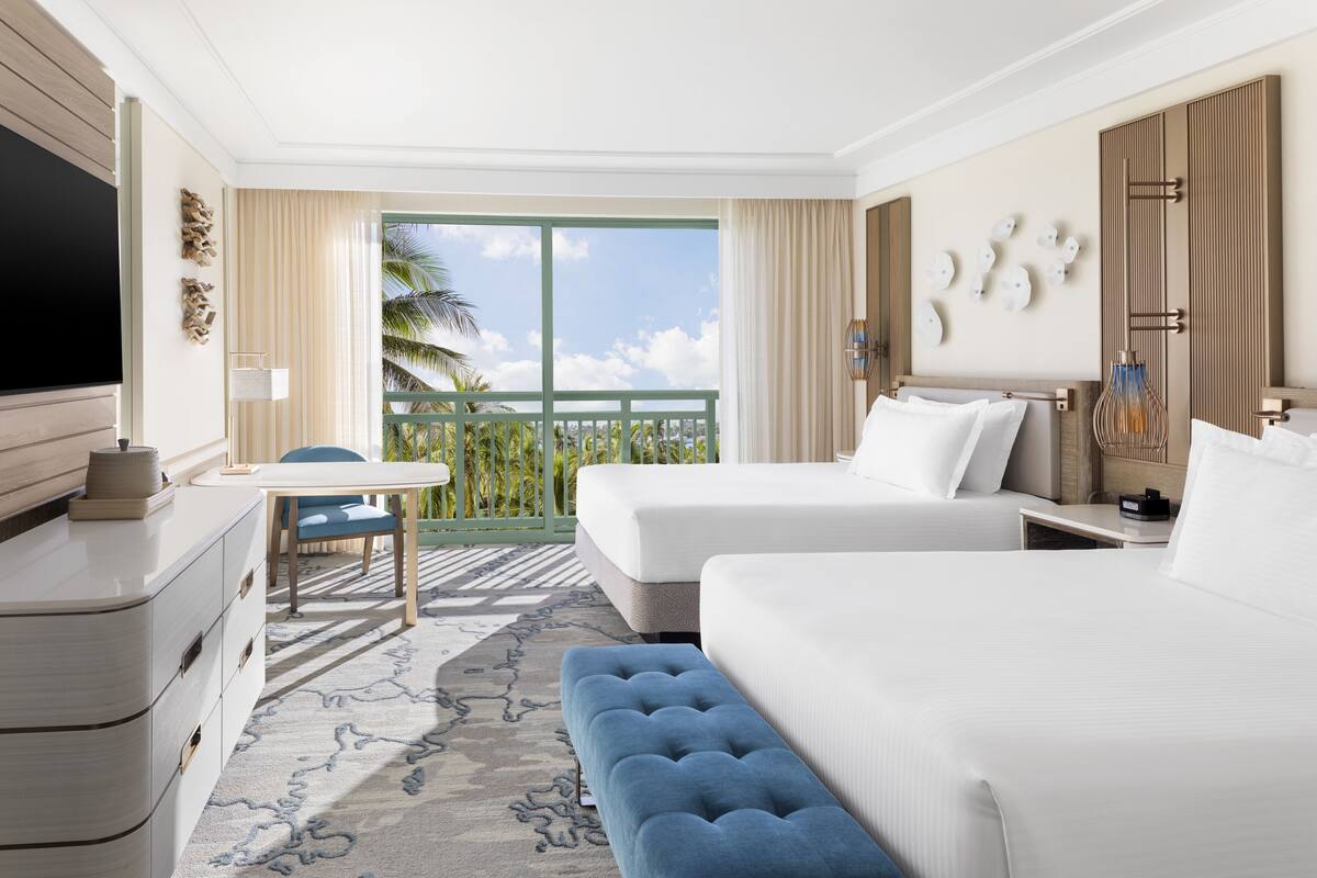 A hotel room with two beds, a TV, a desk, and a balcony with a view of palm trees.