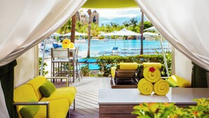 A poolside area with yellow lounge chairs, a table, and a view of a pool and palm trees. - The Royal at Atlantis (Paradise Island)