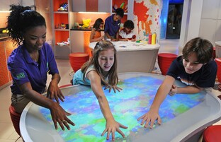 Un grupo de niños interactuando con una mesa colorida e interactiva en una habitación luminosa con paredes anaranjadas y blancas.