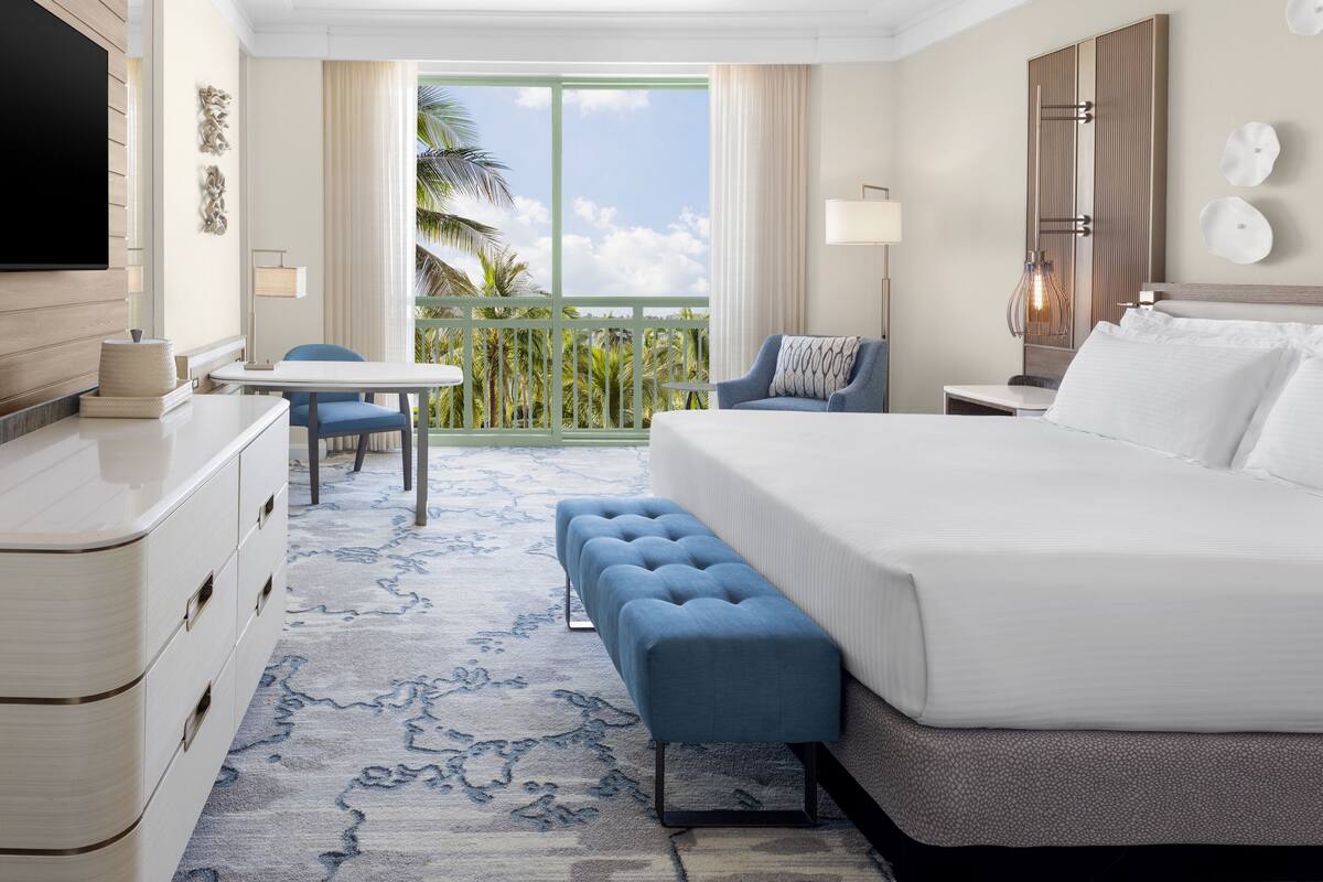 A hotel room with a large bed, a desk with a chair, a small table, a bench, a TV, and a balcony with a view of palm trees.