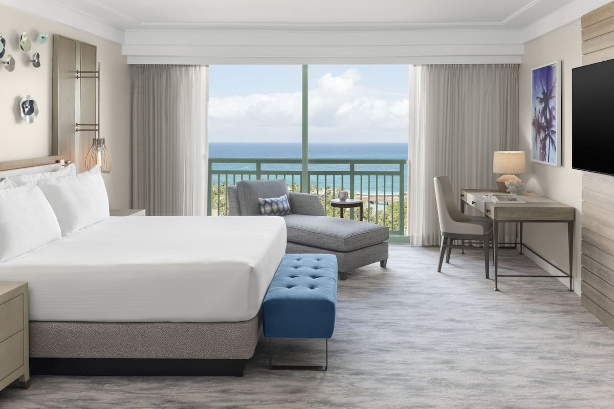 A hotel room with a large bed, a desk, a chair, a small table, and a view of the ocean.