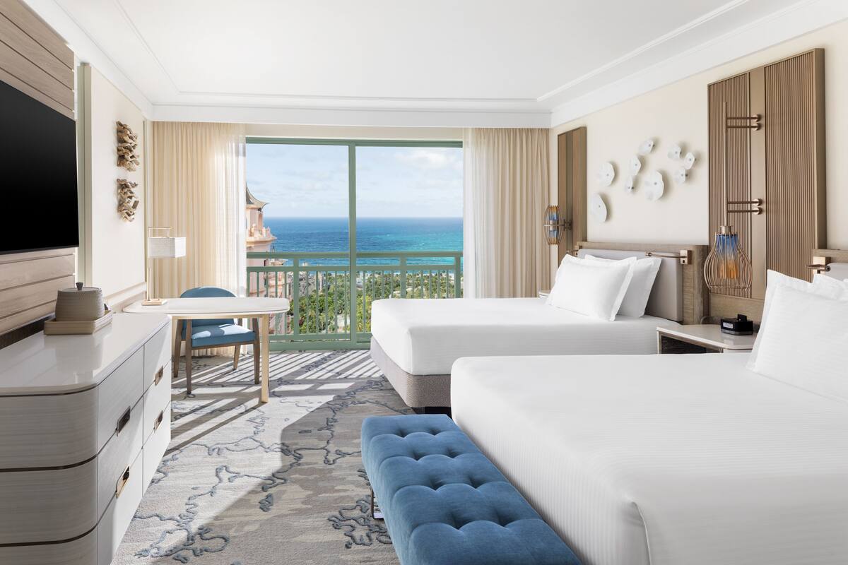 A hotel room with two beds, a TV, a desk, and a balcony with a view of the ocean.