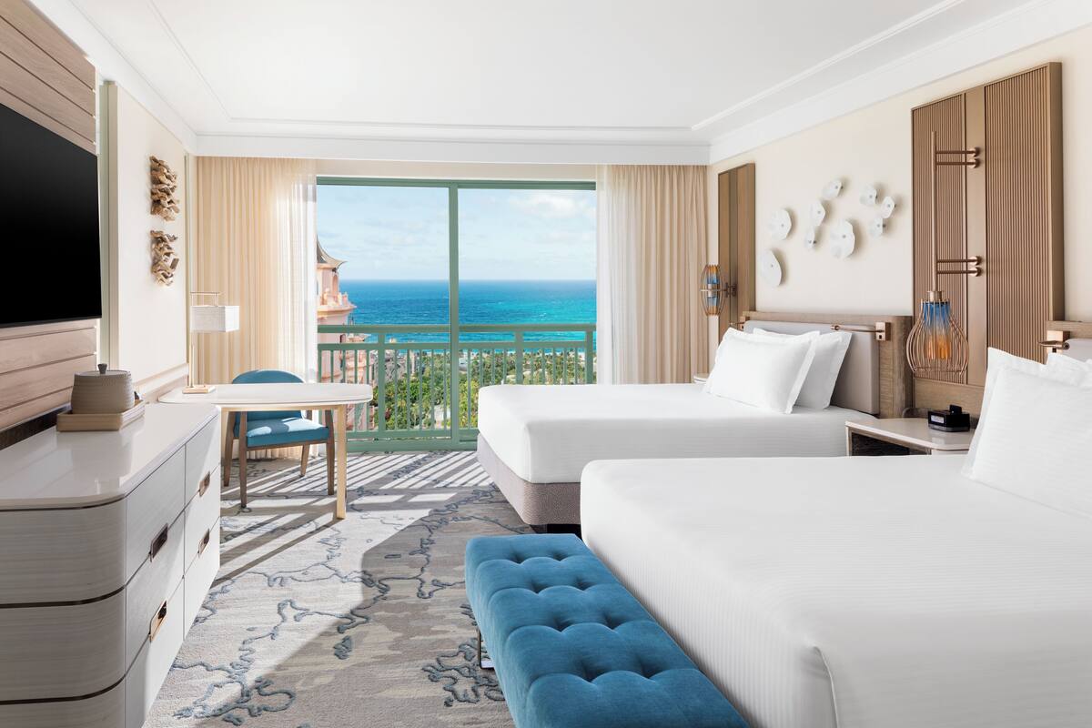 A hotel room with two beds, a TV, a desk, and a balcony with a view of the ocean.