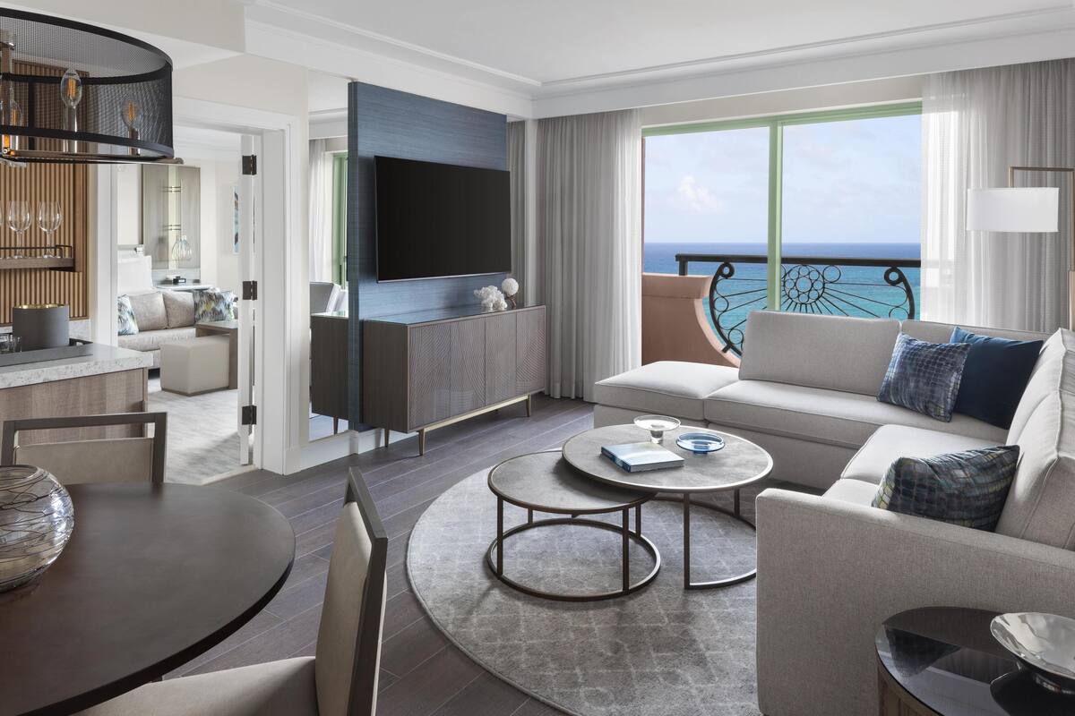 A modern hotel room with a large TV, a sofa, a dining table, and a view of the ocean.