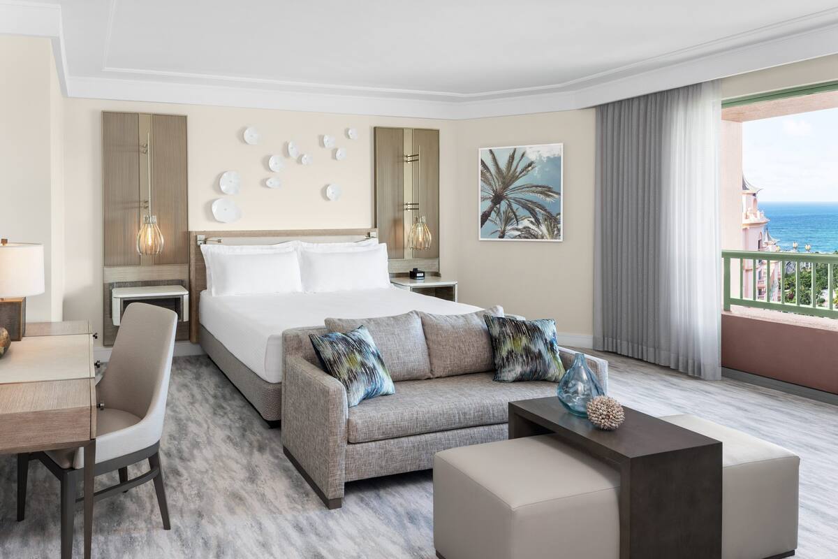 A hotel room with a bed, a sofa, a desk, and a balcony with a view of the sea.