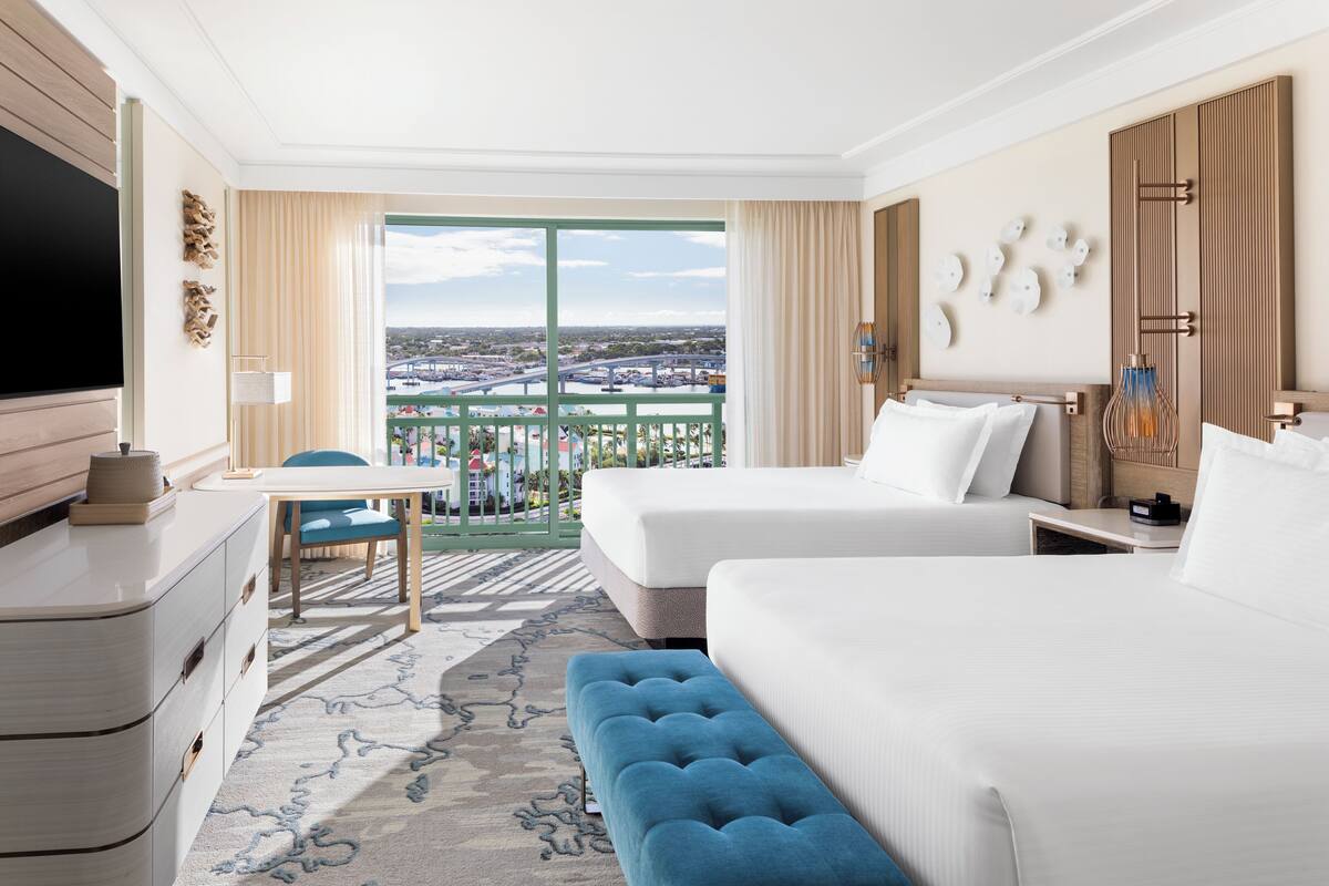 A hotel room with two beds, a TV, a desk, and a balcony with a city view.