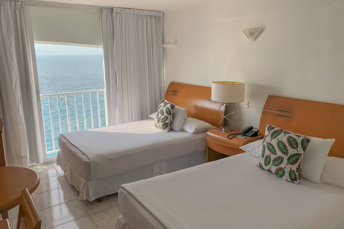 Een hotelkamer met twee bedden, een nachtkastje met een lamp, een telefoon en uitzicht op zee.