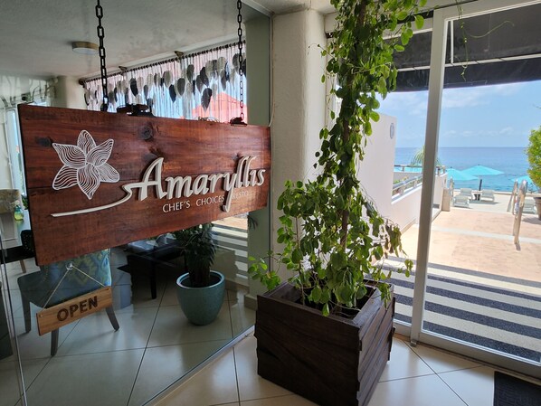 Un letrero de madera con la leyenda 'Amaryllis Chef's Choices' y 'RESTAURANTE', una planta en maceta y vistas al océano a través de un ventanal.