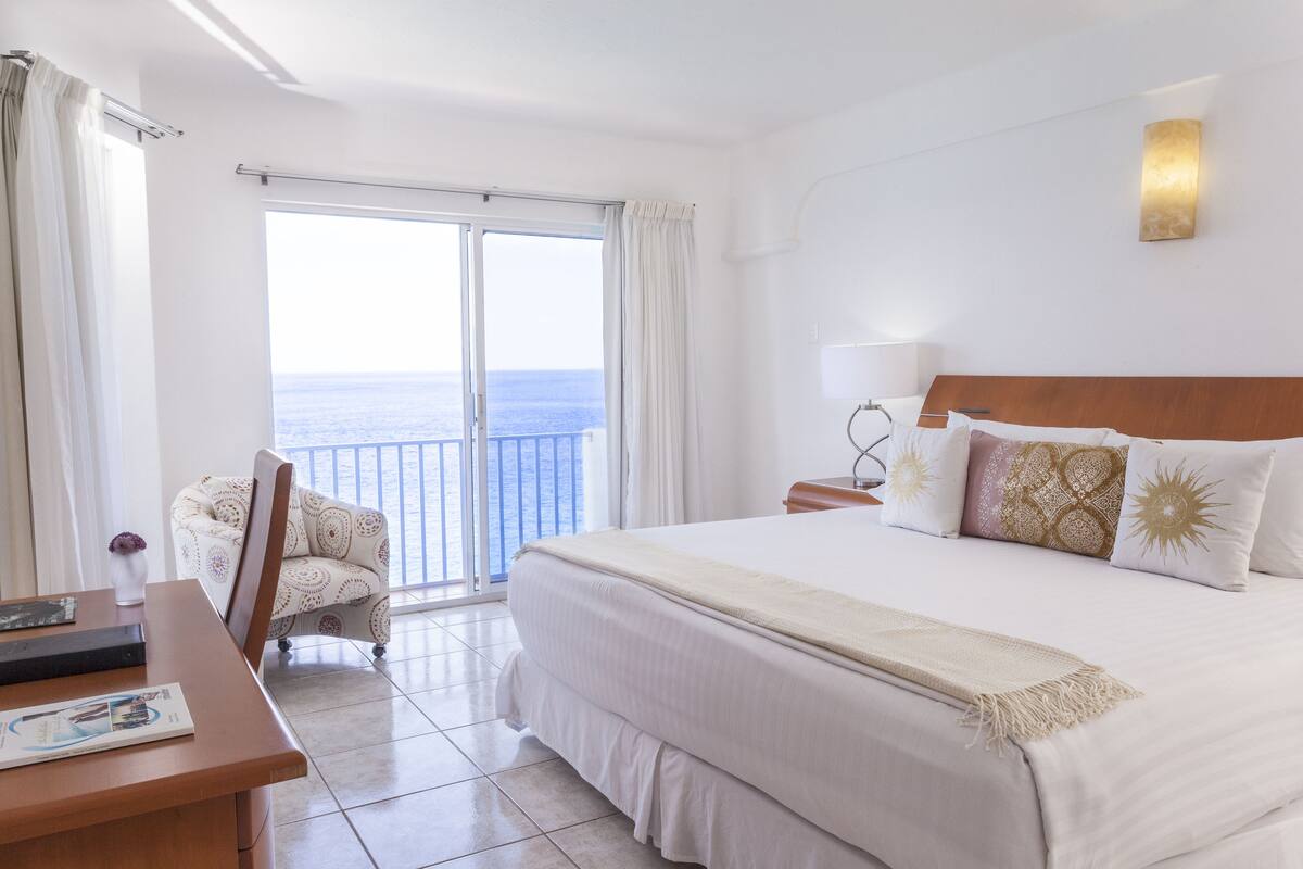 Een hotelkamer met een groot bed, een bureau, een stoel en uitzicht op zee door een schuifdeur.