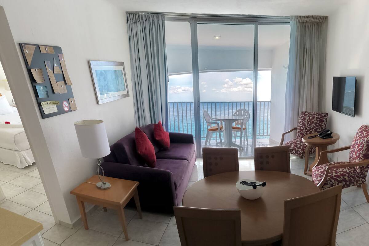 Een moderne woonkamer met een paarse bank, een eettafel met stoelen, een aan de muur bevestigde televisie en een balkon met uitzicht op zee.