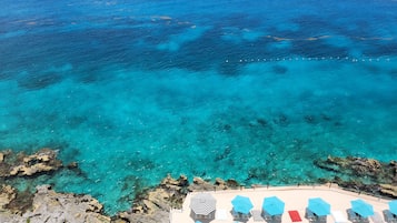 A beachside view with clear turquoise waters, rocky coastline, and a row of sun loungers with umbrellas.