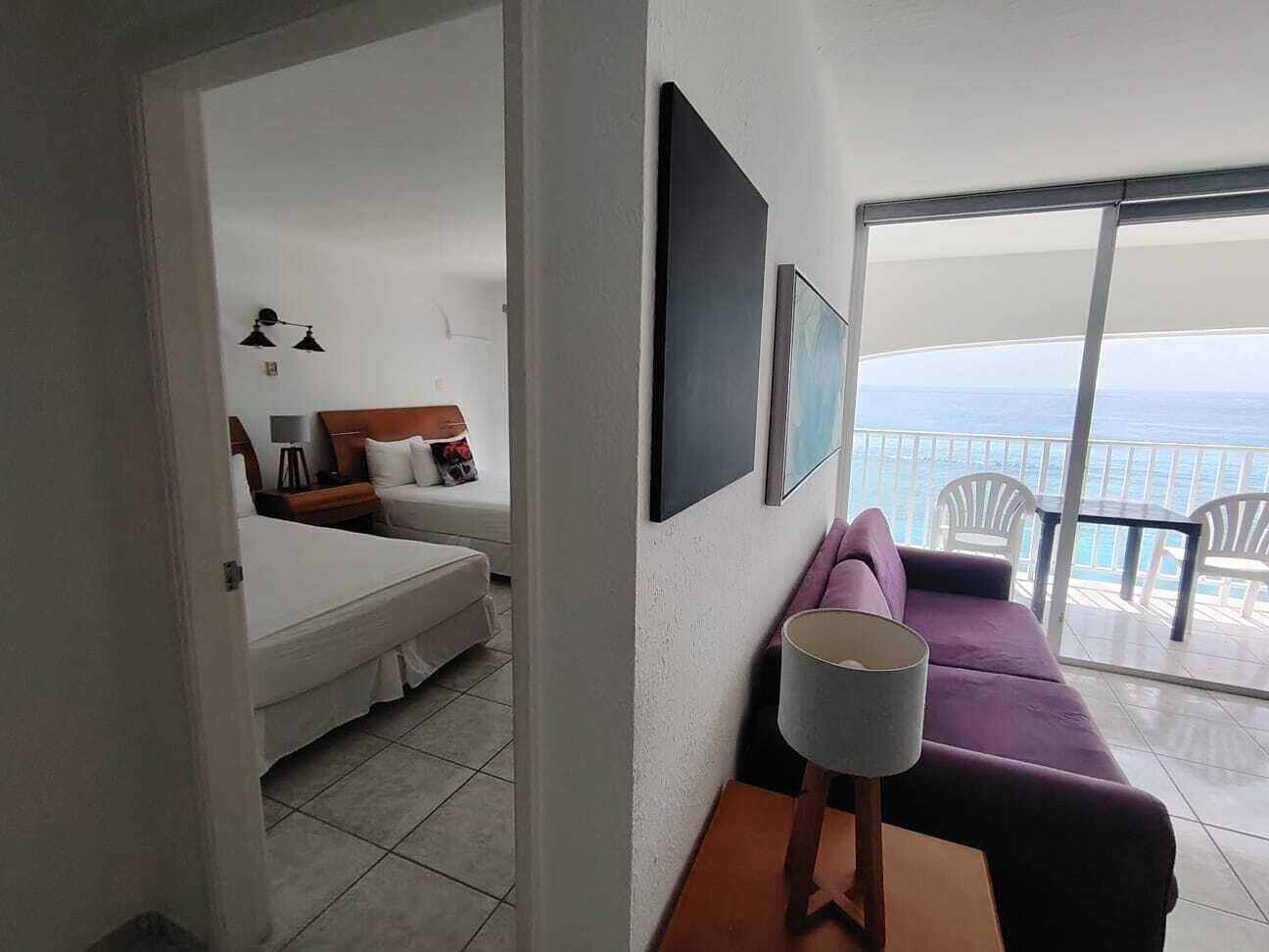 A hotel room with a bed, a purple sofa, a lamp, and a balcony with a view of the sea.
