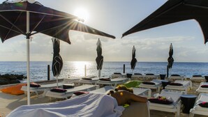 Un espace de détente au bord de la plage, avec des chaises longues et des parasols, une personne allongée sur une table de massage, et l’océan en arrière-plan.