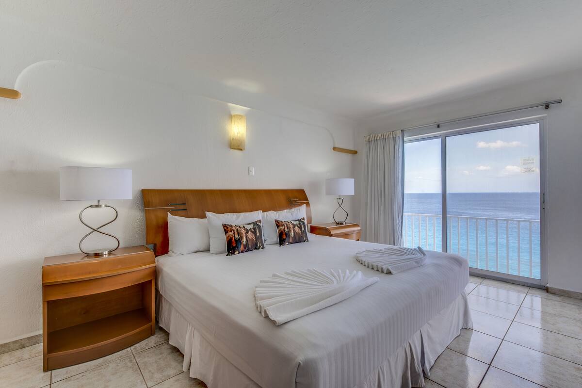 Een hotelkamer met een groot bed, twee nachtkastjes, een bijzettafeltje, uitzicht op de oceaan en een plafondlamp.