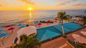 Un espace piscine aménagé dans un complexe hôtelier, avec des chaises longues et des parasols, offrant une vue sur l’océan au coucher du soleil.