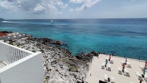 Une vue sur la côte, avec un rivage rocheux, une plage de sable dotée de chaises longues et un voilier à l’horizon.