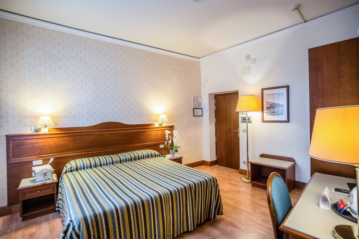 Hotellihuone, jossa on parivuode, yöpöydät, lamppuvarustettu työpöytä, tuoli ja puinen ovi.