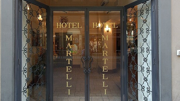 De ingang van Hotel Martelli, met een grote glazen gevel en decoratief ijzerwerk.