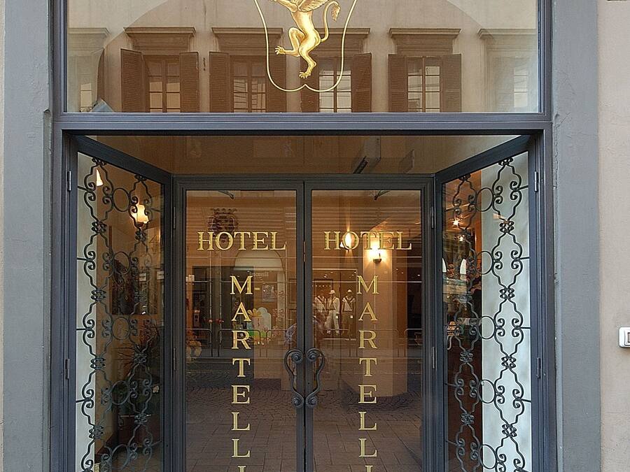 La entrada del Hotel Martelli, con un amplio frente de vidrio y detalles de hierro decorativo.