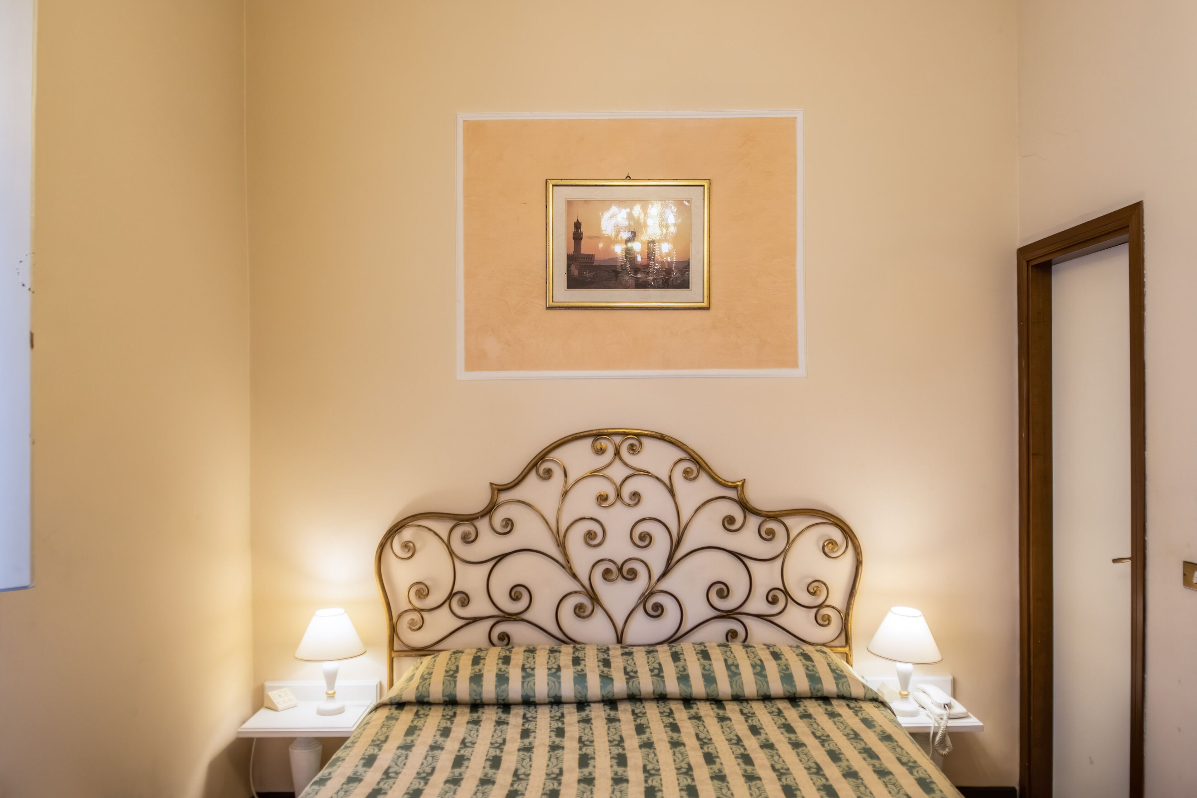 Habitación de hotel con cama, dos mesitas de noche con lámparas, un cuadro enmarcado en la pared y una puerta.