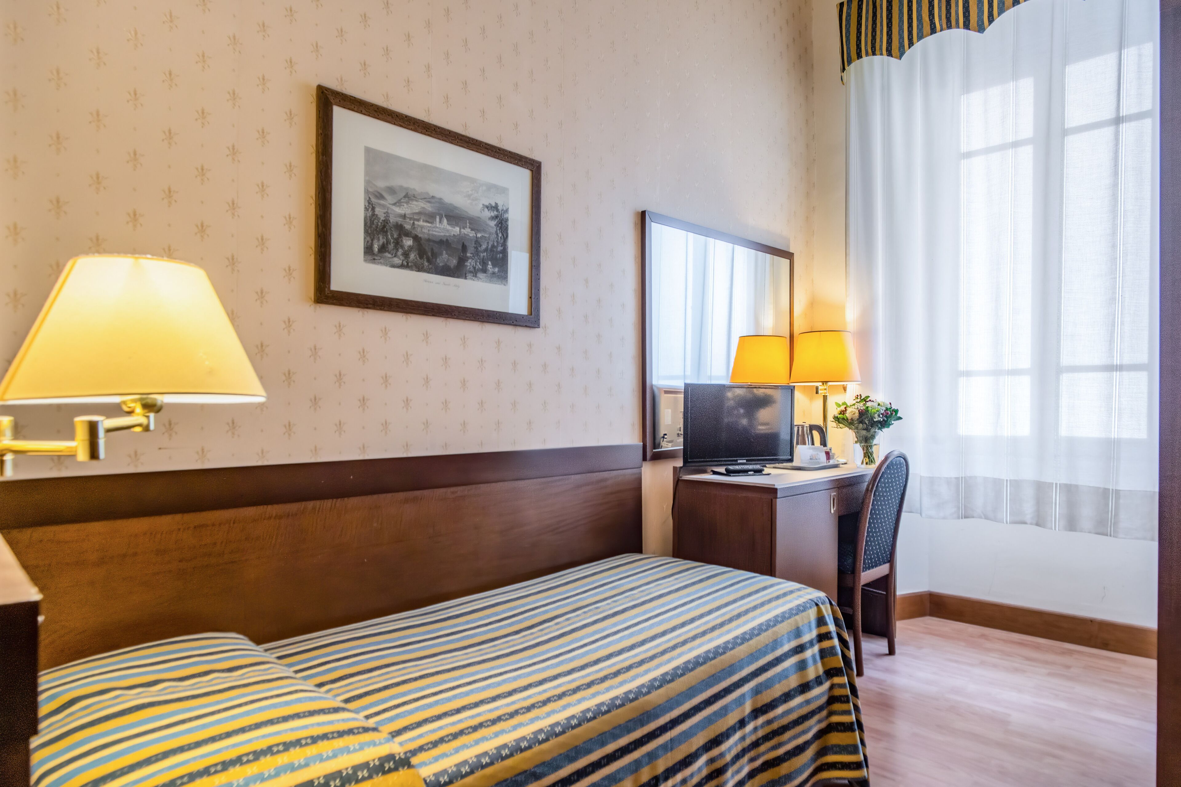 Une chambre d’hôtel avec un lit, un bureau avec un ordinateur, une chaise, une lampe et une fenêtre avec des rideaux.