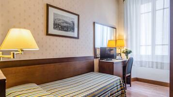 Une chambre d’hôtel avec un lit, un bureau avec un ordinateur, une chaise, une lampe et une fenêtre avec des rideaux.