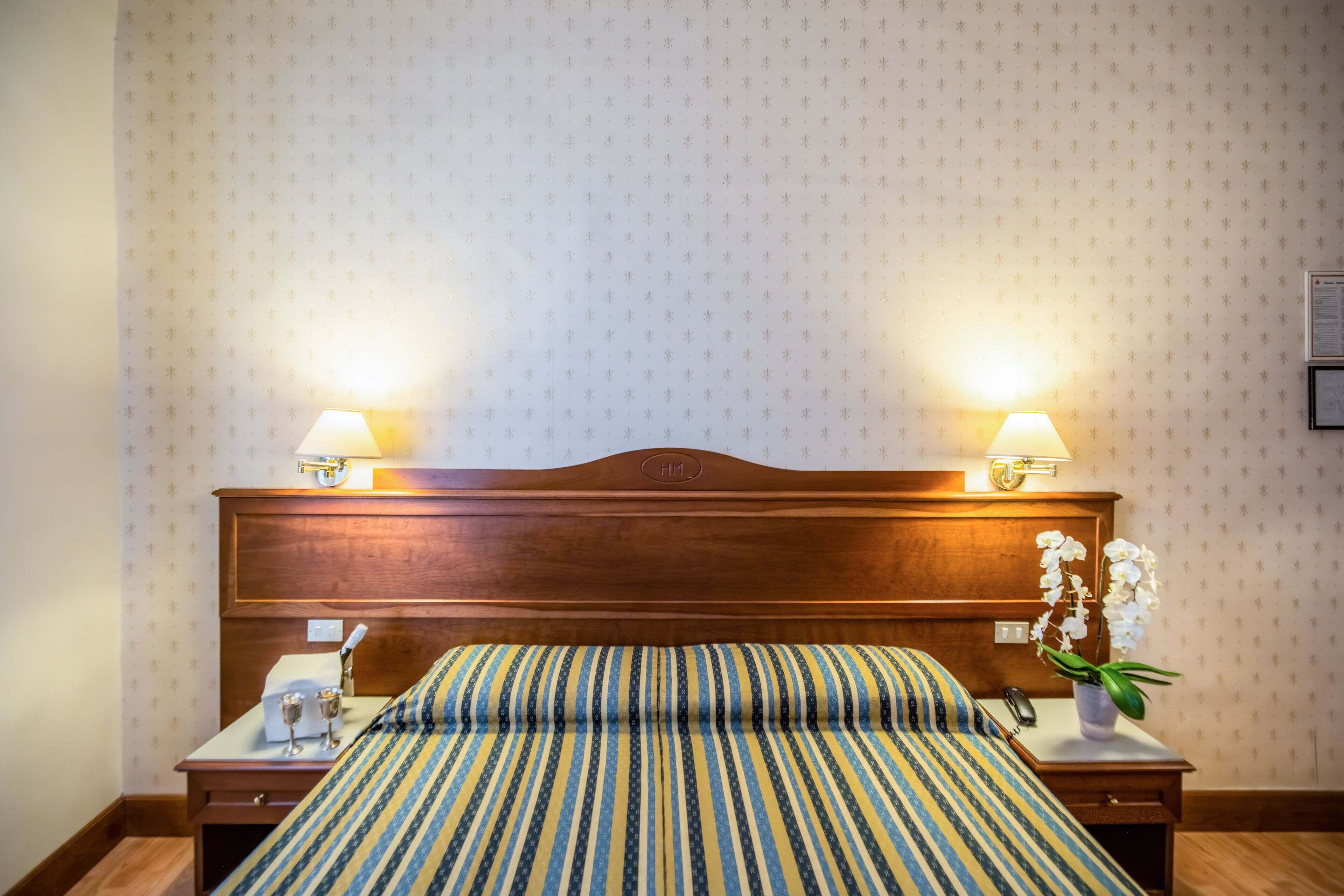 A hotel room with a wooden headboard, two bedside lamps, a striped bedspread, a bedside table with a telephone, and a vase with an orchid.