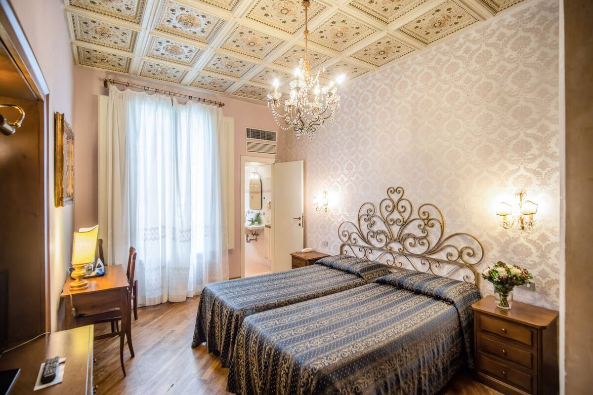 Hotellihuone, jossa on kaksi sänkyä, kirjoituspöytä lampun kanssa, kattokruunu ja peili.
