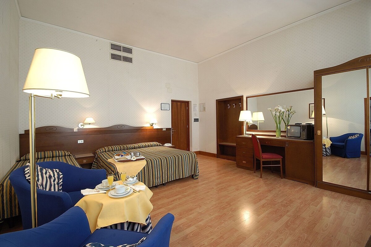 Habitación de hotel con cama, escritorio, silla, mesa con productos para el desayuno, lámpara y espejo.