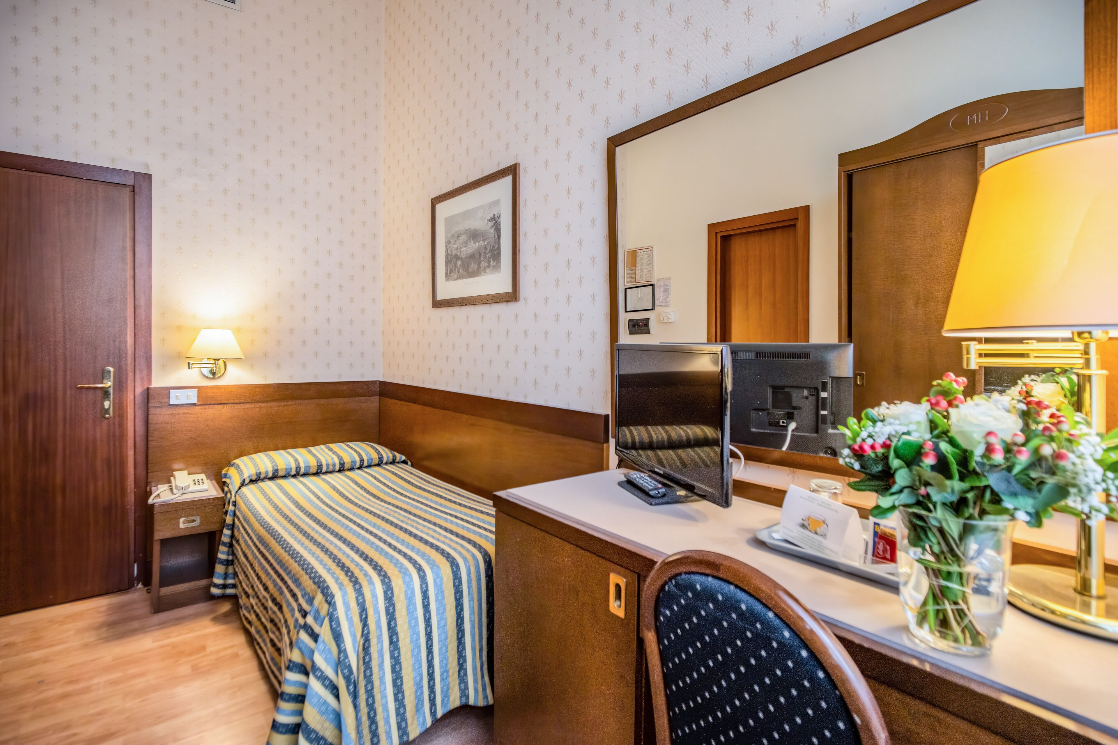 Una camera d'albergo con un letto, una scrivania con un computer, un telefono, uno specchio e un vaso con fiori.